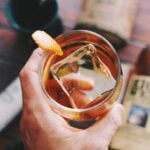 A hand holding a clear cup full of whiskey with a giant ice cube over a blue background