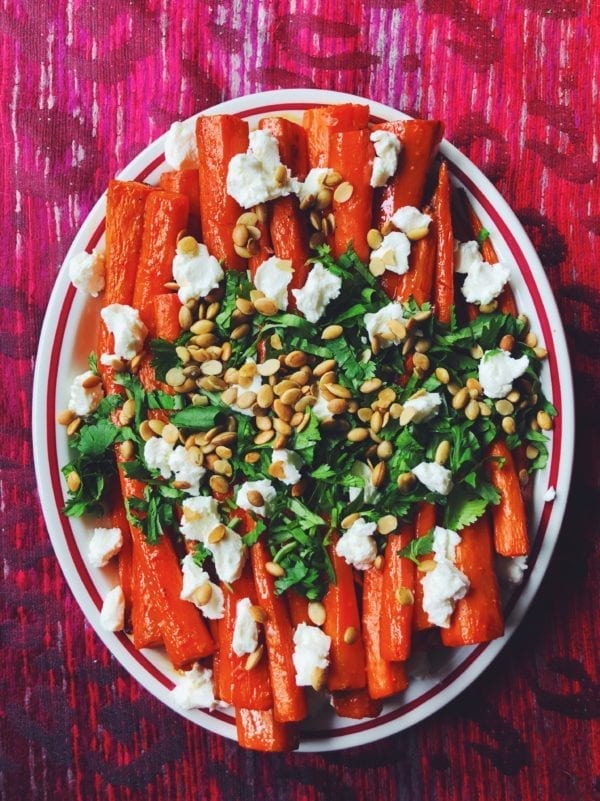 Honey Sriracha Roasted Carrots with Goat Cheese, Pepitas + Cilantro on a white plate and a pink background