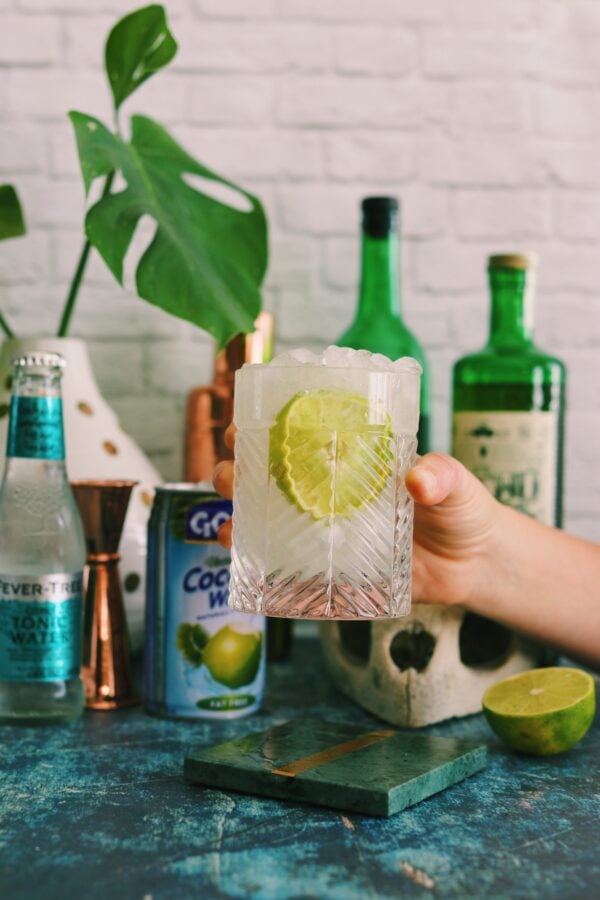 a hand holding a mezcal cocktail in a clear glass with a big lime wedge in it. The background has bottles and a turtle skull with fresh palm leaves