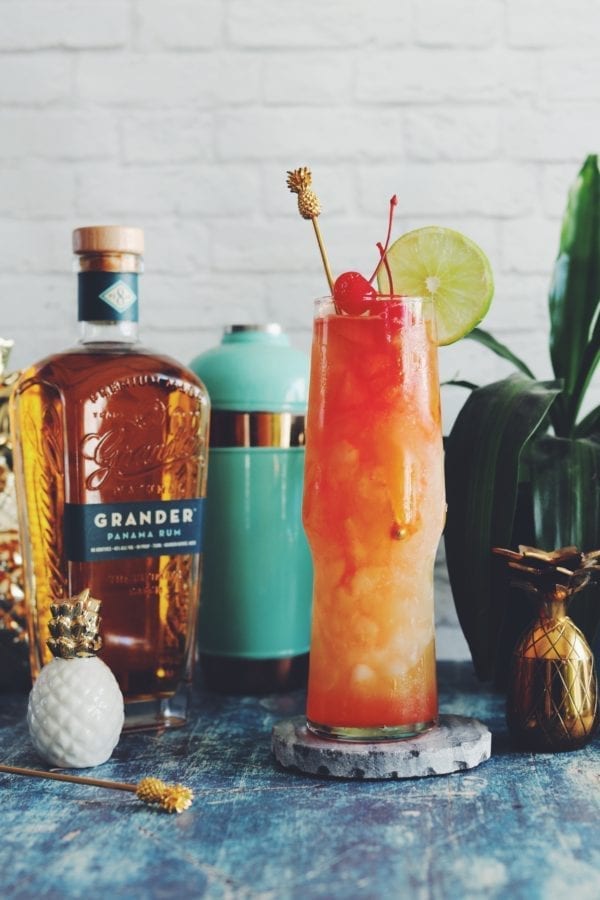 A tall orange and red drink against a white background with a blue table with cocktail making ingredients in the back