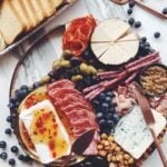 a wooden cheese plate full of meats, pickles, and fresh fruit on a white marble counter top