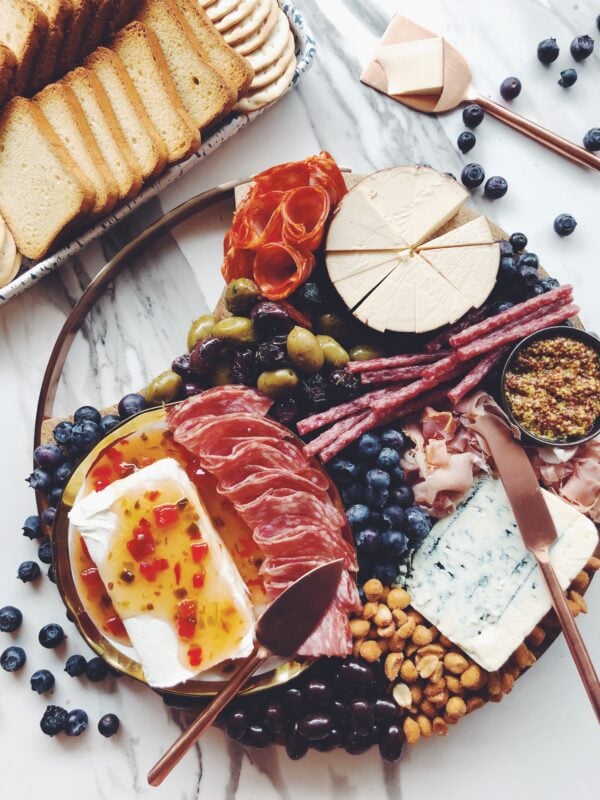 a wooden cheese plate full of meats, pickles, and fresh fruit on a white marble counter top