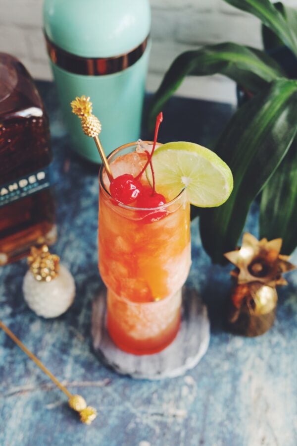 A birds eye view of a tall orange and red drink against a white background with a blue table with cocktail making ingredients in the back