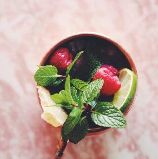 one copper mule cup filled with hfresh mint leaves, lime, raspberries and candied ginger on a pink marble background