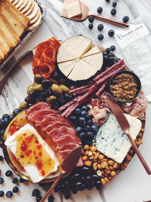 a wooden cheese plate full of meats, pickles, and fresh fruit on a white marble counter top with a receipt in a corner.