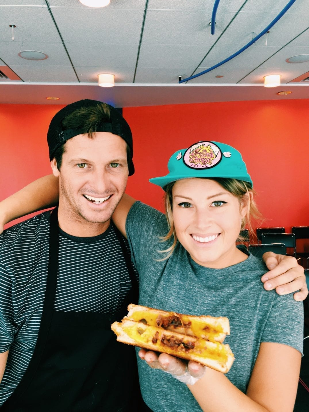 MacKenzie Smith and Jeremy Johnston holding a grilled cheese with a red background