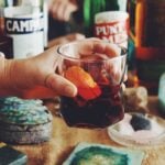 a hand holding a clear glass full of redish brown liquor with an orange slice in it - various bottles in the background