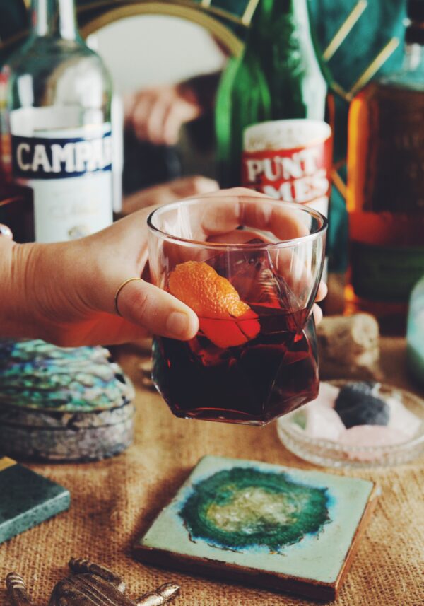 a hand holding a clear glass full of redish brown liquor with an orange slice in it - various bottles in the background