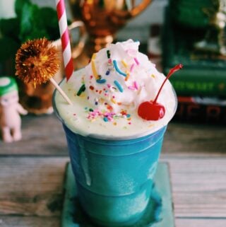 a shiny aqua cup filled with a green milkshake with rainbow sprinkles, whipped cream and a cherry on top