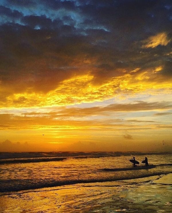 a yellow sunrise with two surfers walking out into the water