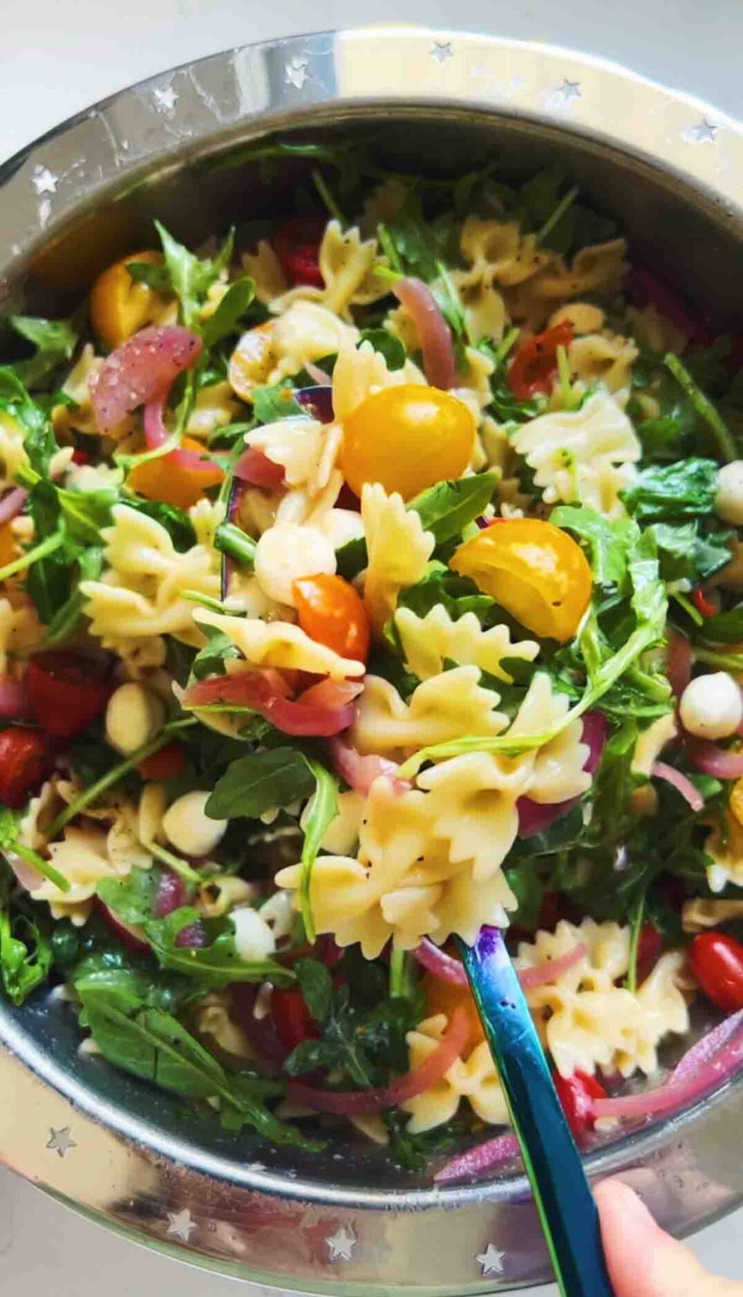 a blue spoon holding caprese pasta salad over a silver bowl.