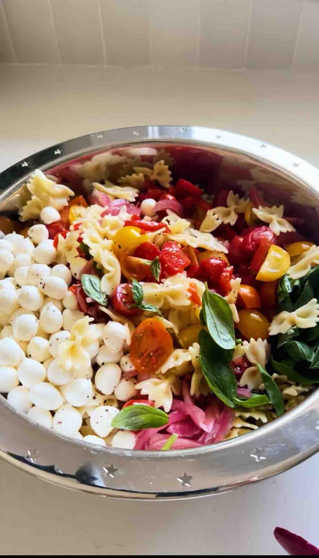 a silver bowl filled with caprese pasta salad ingredients on a white countertop.