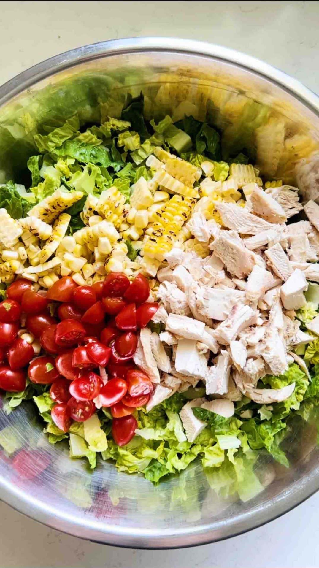 romaine lettuce, rotisserie chicken, tomatoes and grilled corn in a big silver bowl on a white surface.