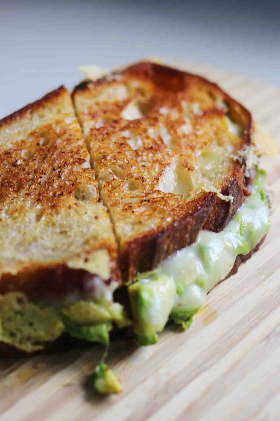 an avocado grilled cheese cut in half on a wooden cutting board.