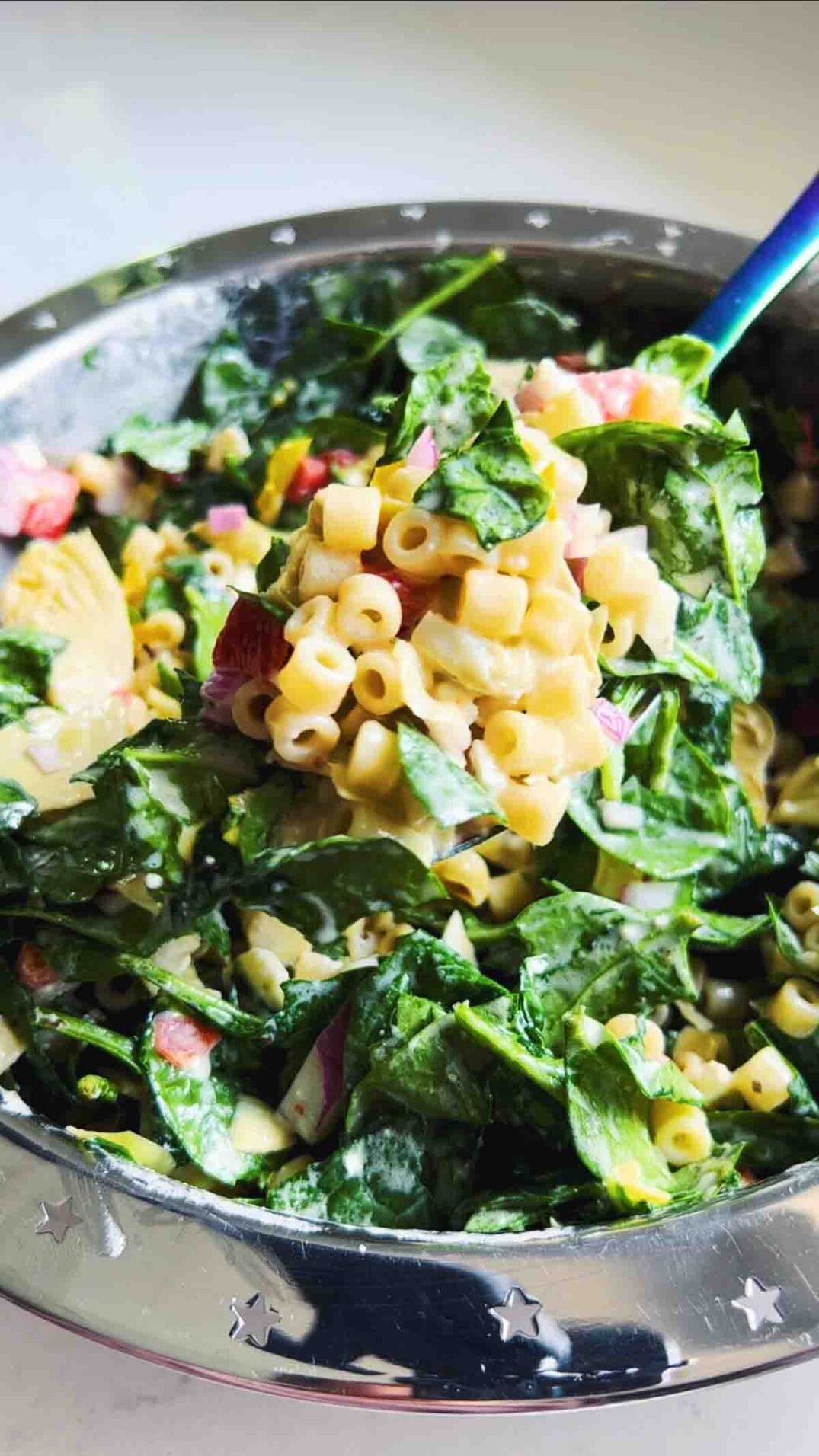 a blue spoon holding spinach artichoke pasta salad over a silver bowl.