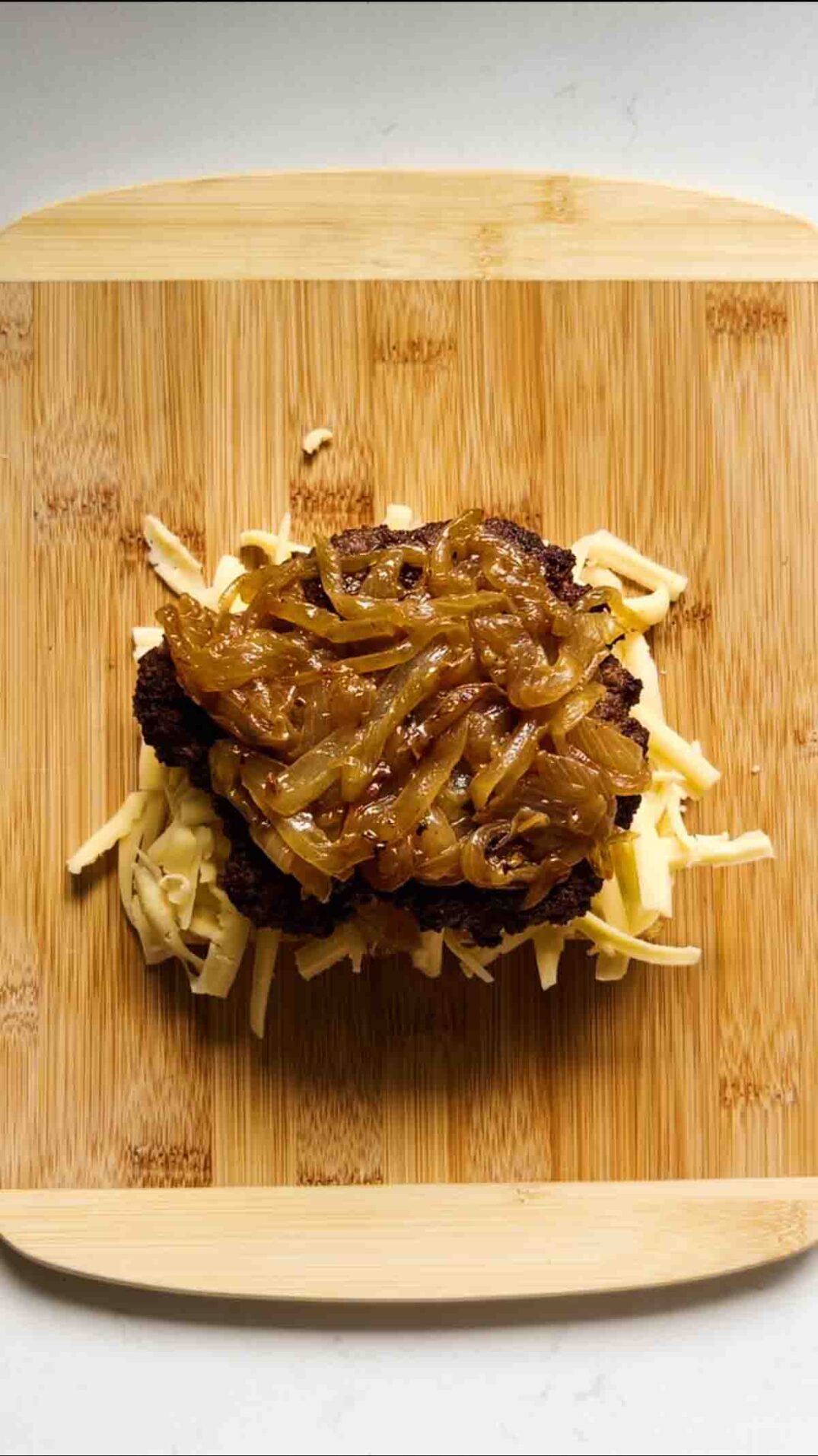 caramelized onions, smash burger patty and shredded cheese on a cutting board.