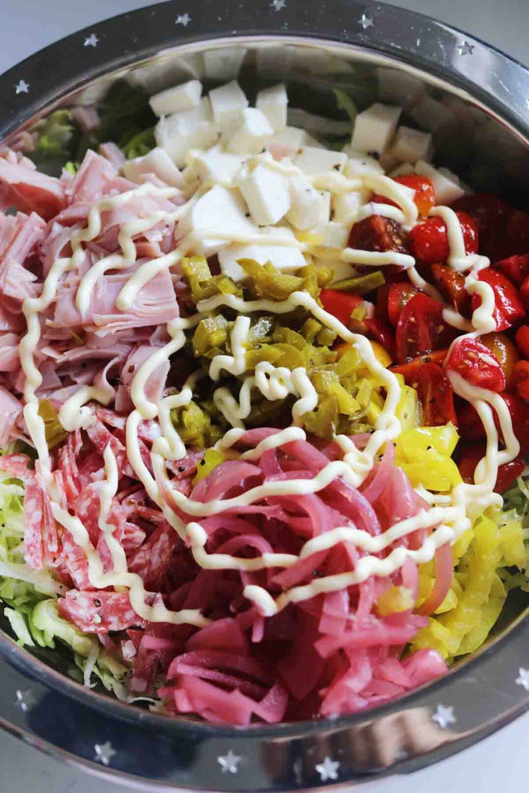 mayo swirled above chopped up colorful veggies, italian cold cuts, and mozzarella in a silver bowl with shredded lettuce.