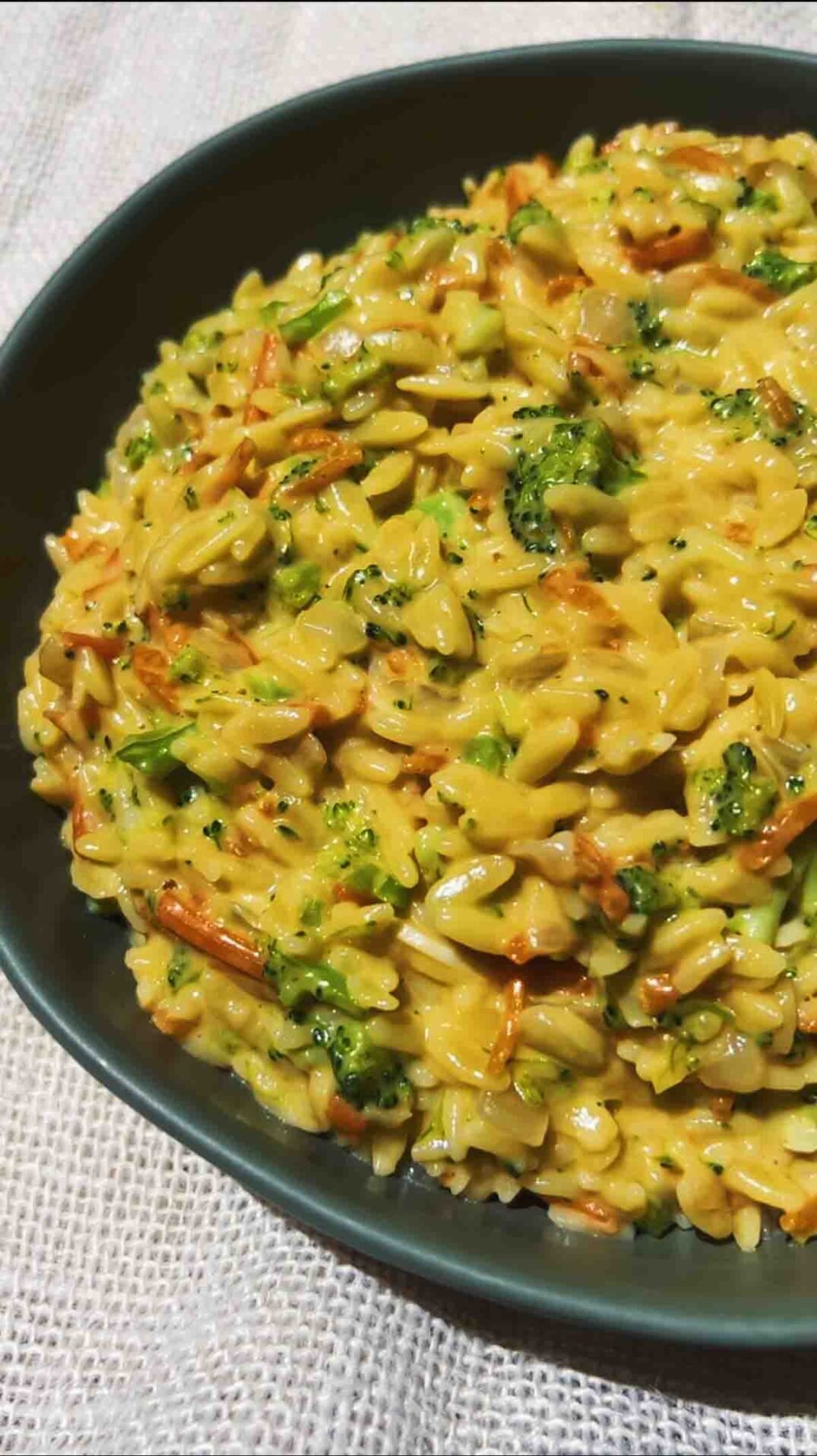 cheesy orange orzo in a green bowl with bits of broccoli and carrot in it.