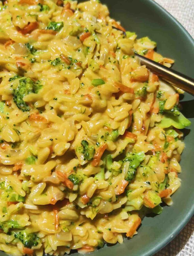 a close up of cheesy orzo with broccoli, a for, and a green bowl on a white tablecloth.