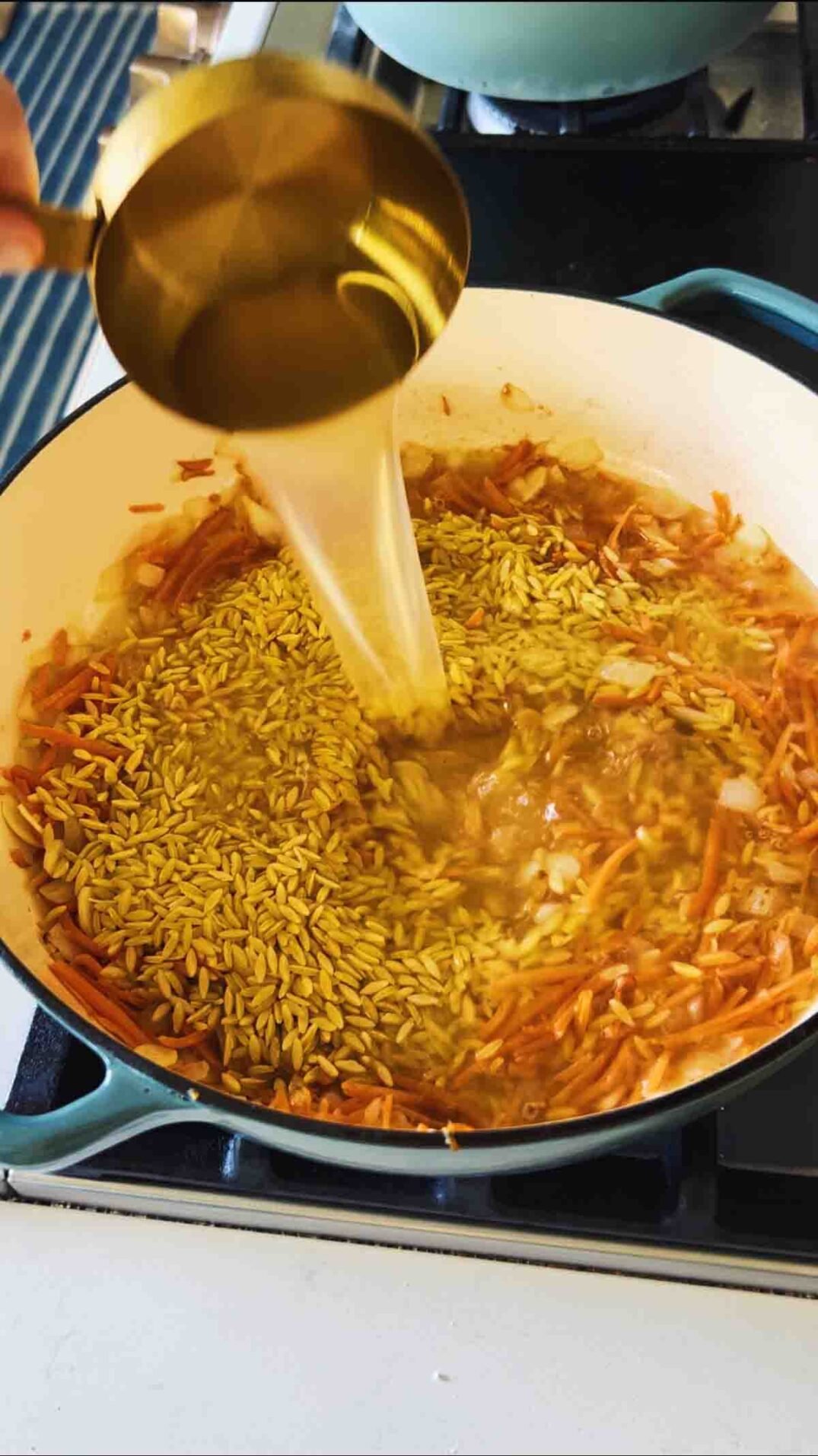 chicken stock being poured into orzo in a blue braising dish on the stovetop.