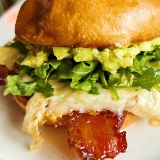 candied bacon, cheesy eggs, arugula, and avocado on a bun on a white plate with a pink rim.