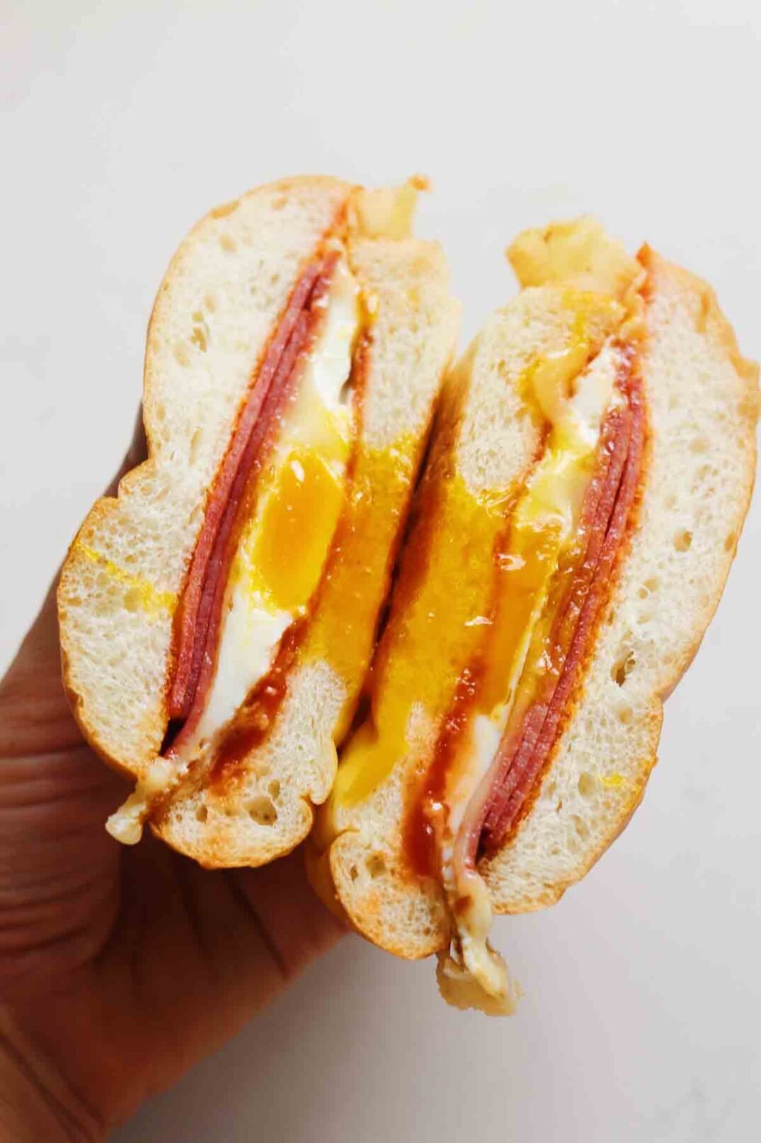 a hand holding a split open pork roll egg and cheese sandwich over a white background.