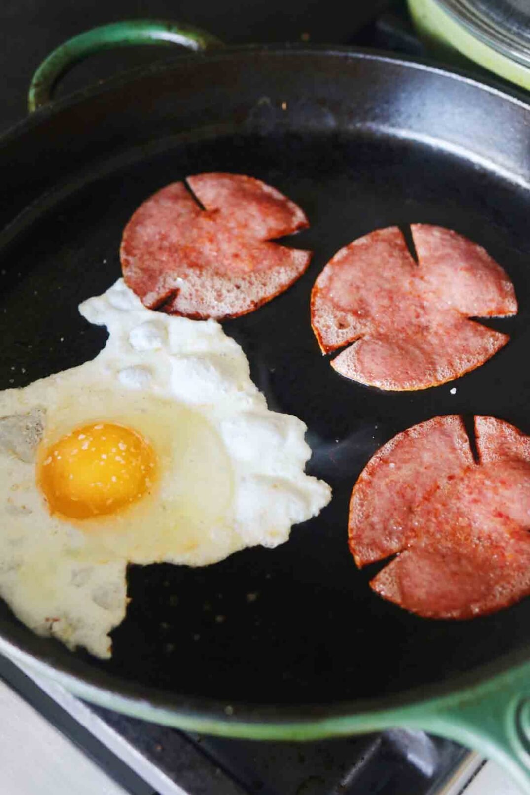 an egg cooking in a green fry pan with pork roll in the background.