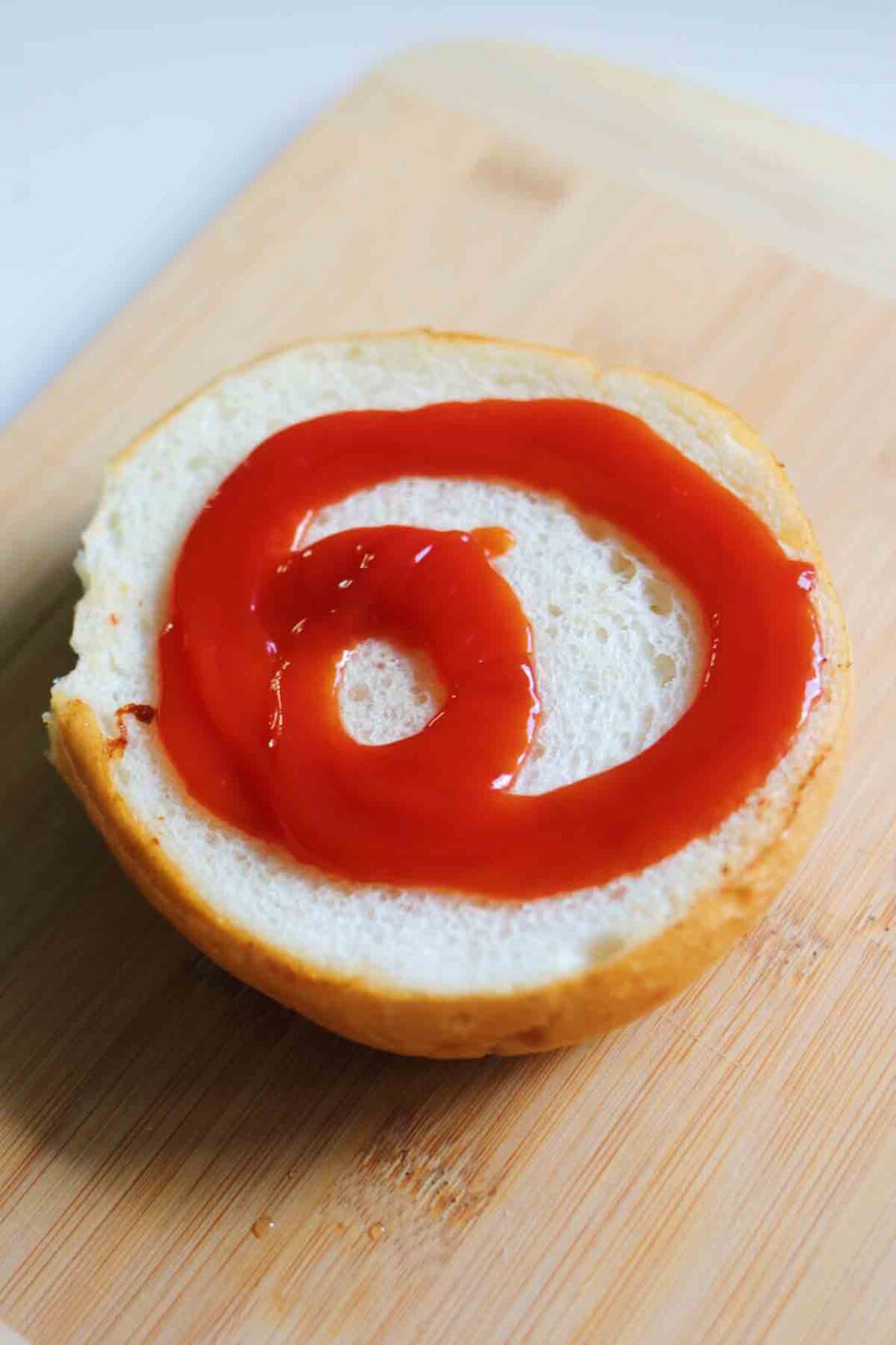 ketchup drizzled on the bottom half of a kaiser bun on a wooden cutting board.