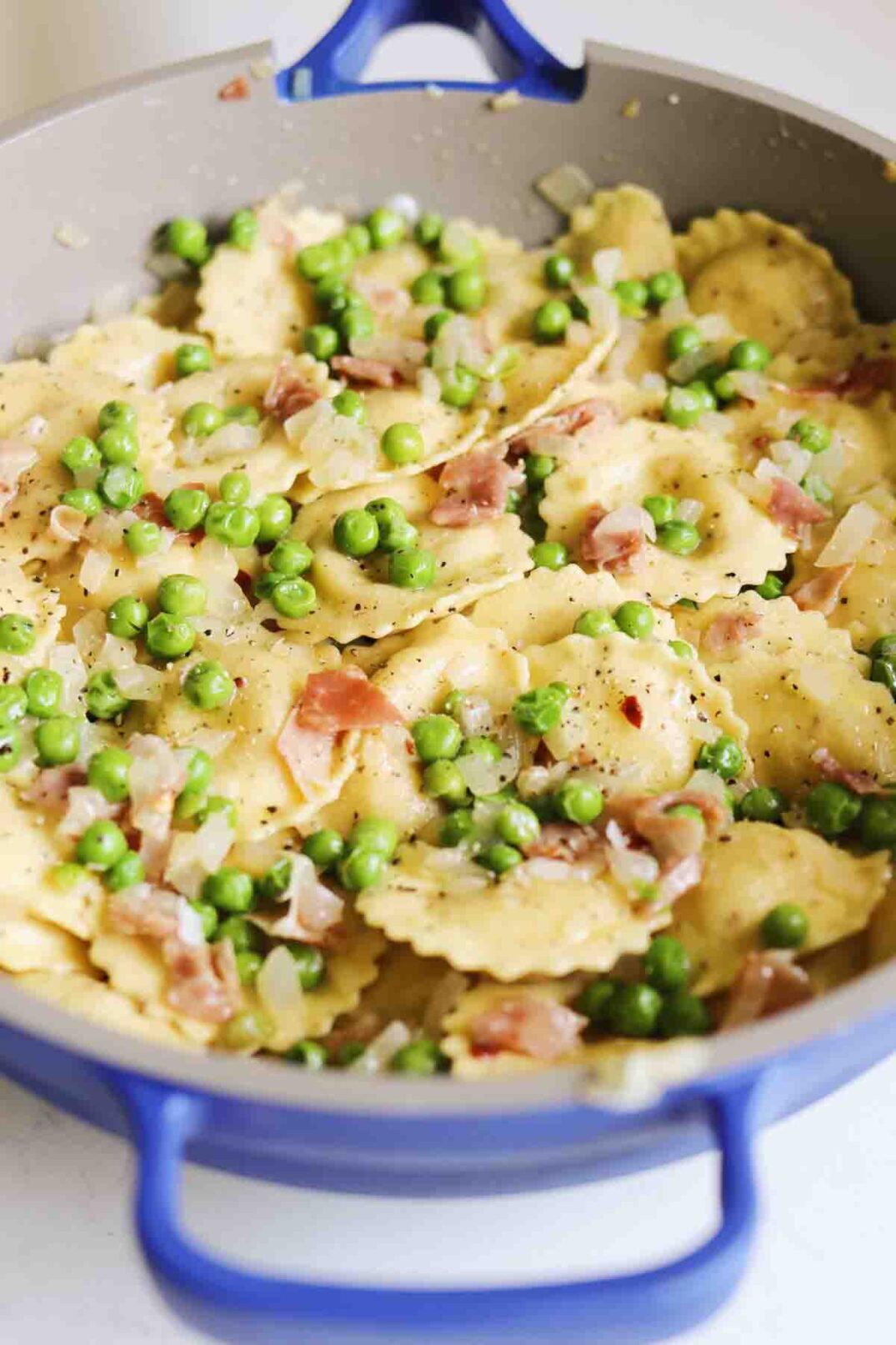 a blue pot filled with tiny green peas, pink crispy prosciutto and cream colored ravioli.
