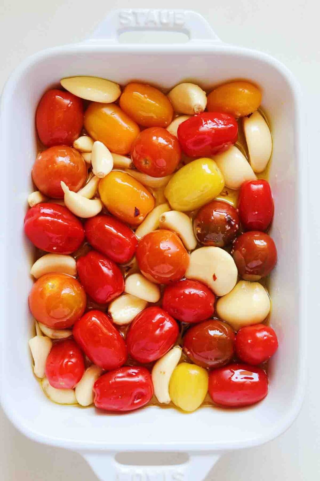 fresh cherry tomatoes drizzled with olive oil with big fresh chunks of garlic in a white baking dish.