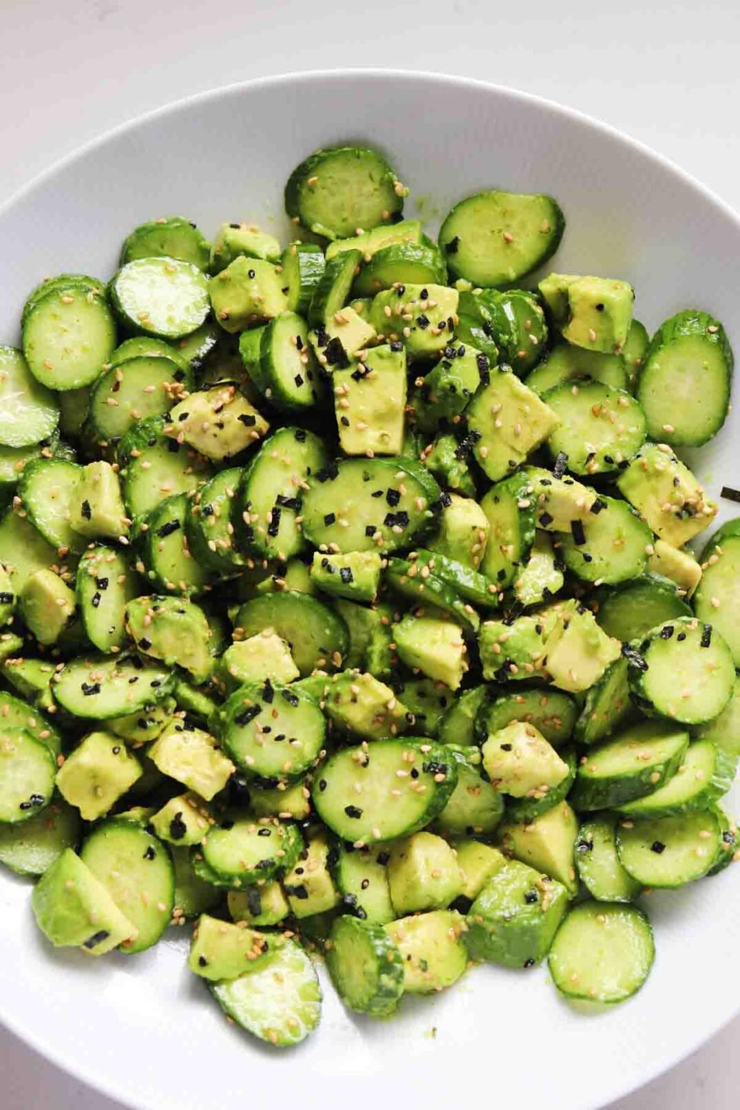cubed avocado, sliced cucumbers and sprinkles of furikake in a white bowl on a white counter.