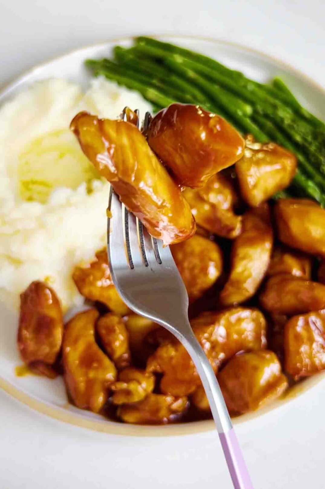a fork holding a piece of honey bourbon chicken over mashed potatoes and air fryer asparagus. 