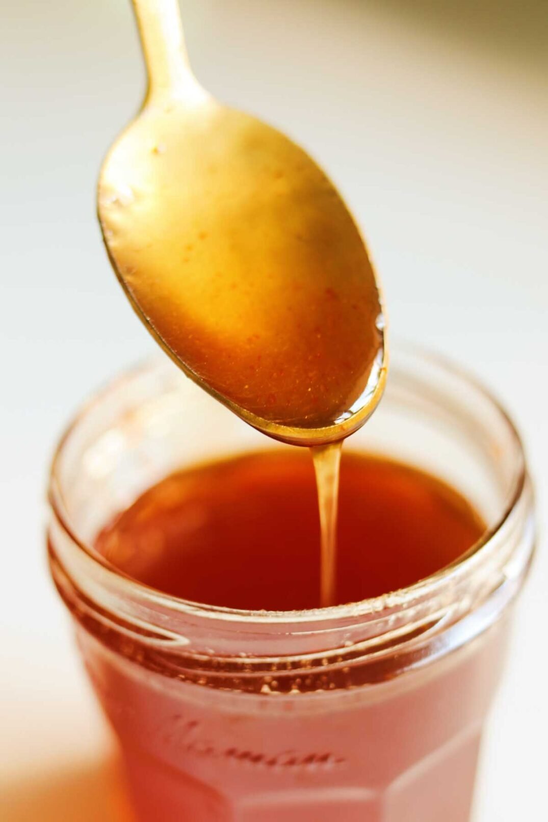 a golden spoon dripping hot honey sauce into a glass jar. 