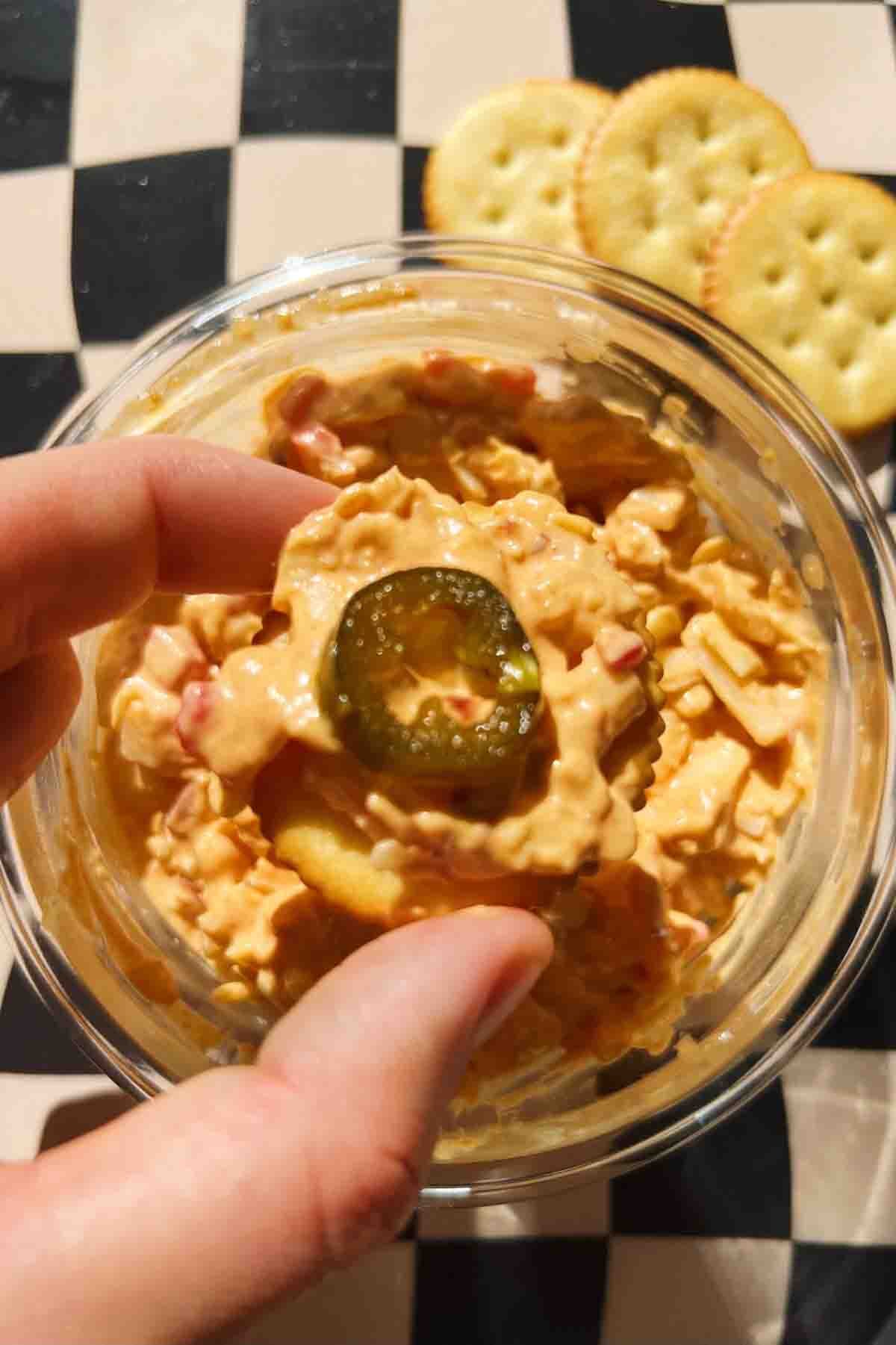 a hand holding a cracker with pimento cheese dip and a candied jalapeno.