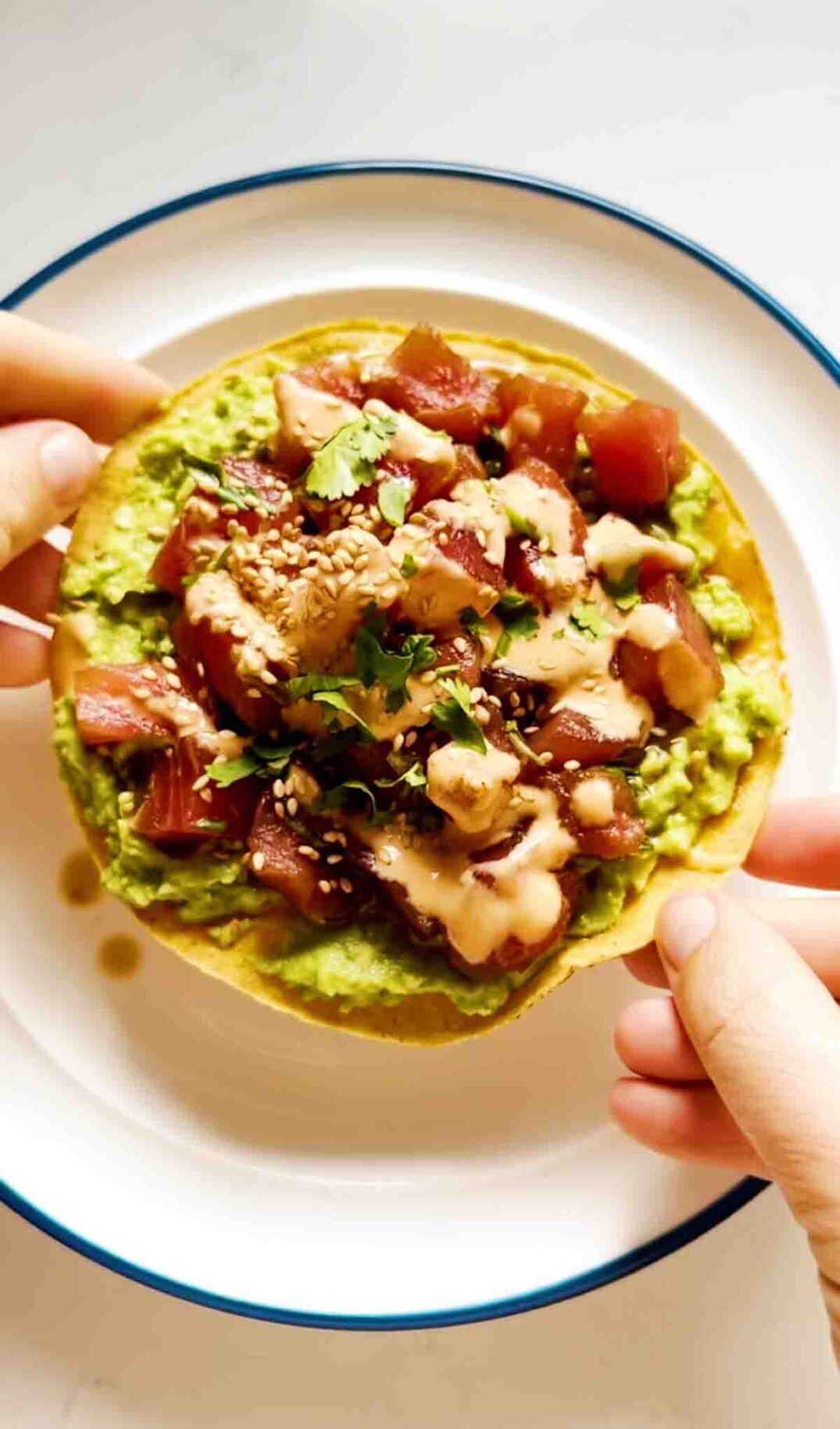 two hands holding a tuna tostada over a white plate. 