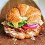 a bright and colorful turkey croissant sandwich on a wooden cutting board.