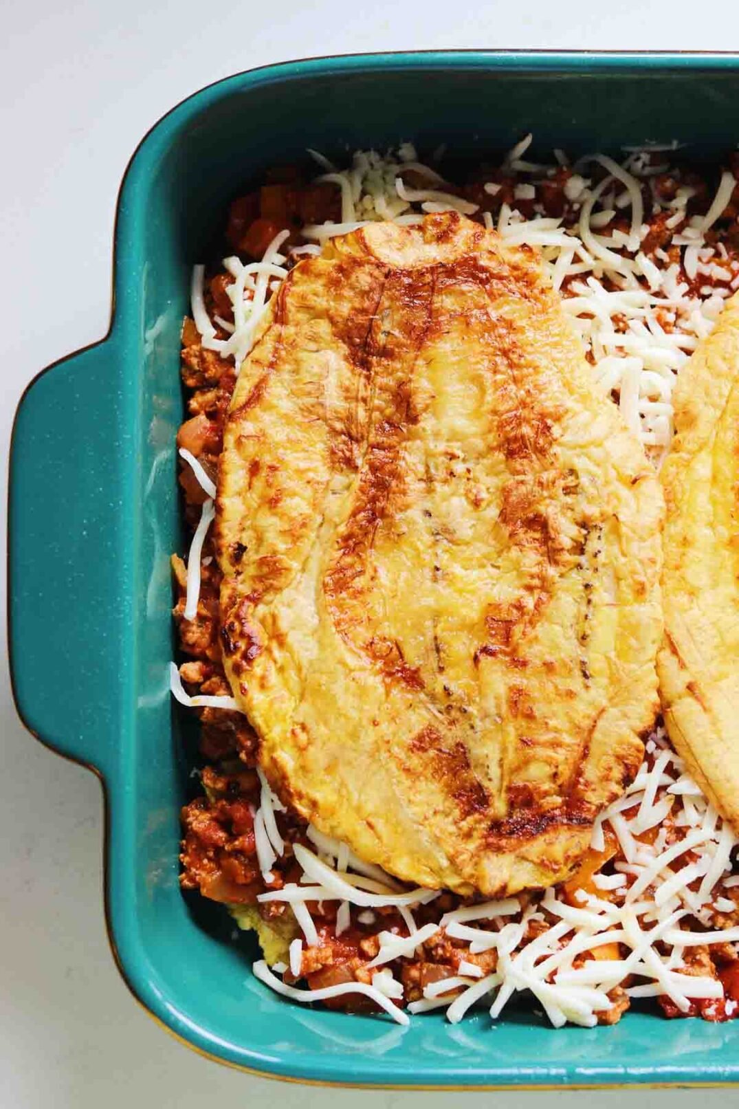 smashed plantains on top of meat and cheese in a blue baking dish. 