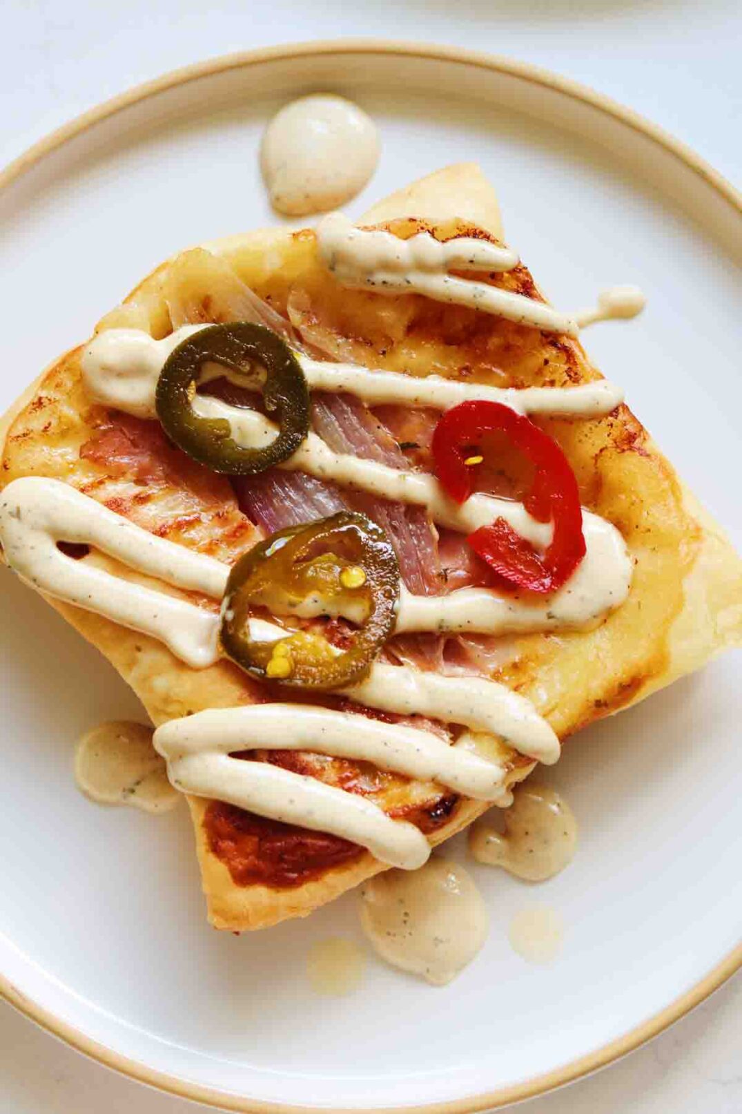 a white plate with upside down puff pastry topped with a drizzle of white sauce and pickled jalapenos.