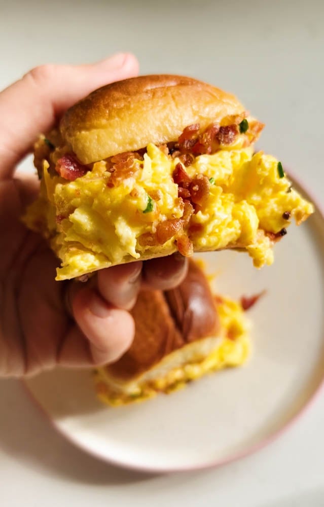 a hand holding a hawaiian roll filled with scrambled eggs, bacon, chives and spicy mayo on a hawaiian bun.