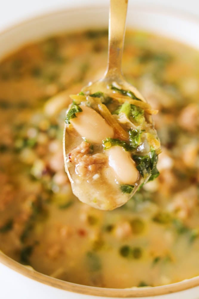 a spoon filled with sausage, kale, and white bean soup. 