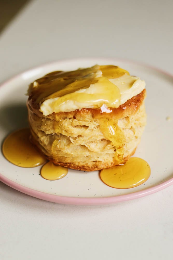a singular honey butter biscuit getting a drizzle of honey on a white plate.