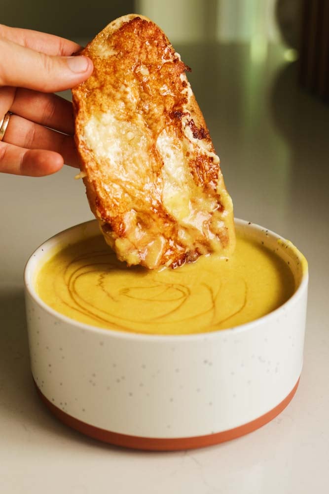 a piece of crispy feta toast being dunked into a bowl of soup. 