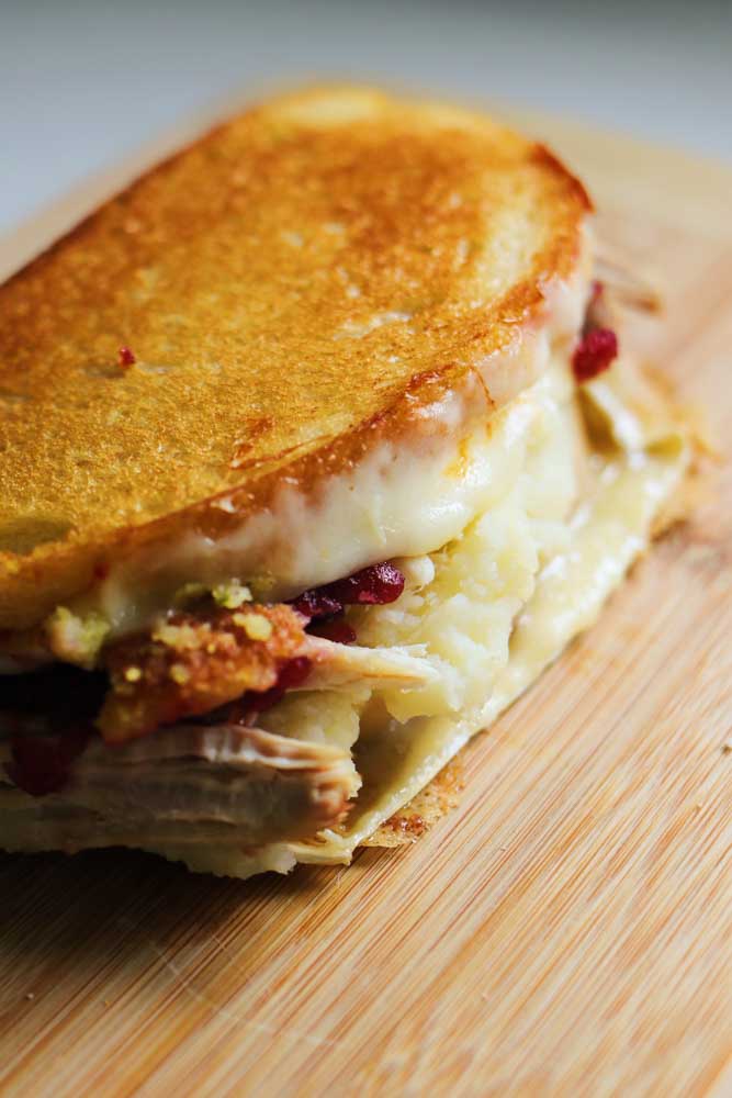 a whole turkey cranberry sandwich sitting on a wooden cutting board with cheese oozing out. 