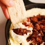 a cracker dipping into a cast iron skillet filled with whipped goat cheese dip with hot honey bacon and dates.