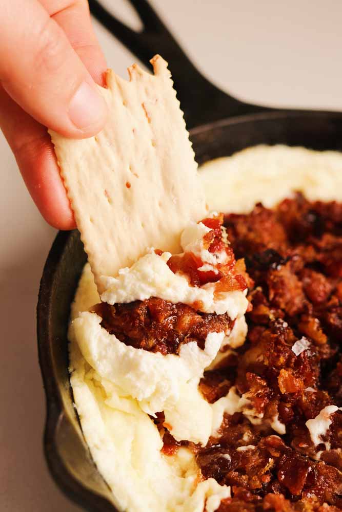 a cracker dipping into a cast iron skillet filled with whipped goat cheese dip with hot honey bacon and dates.