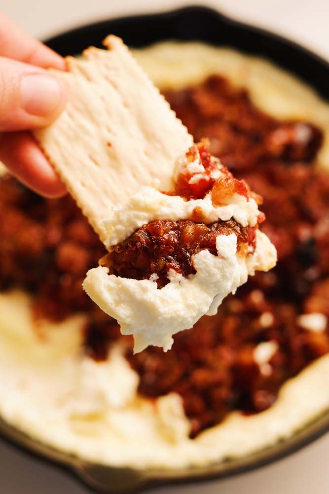 a cracker dipping into whipped goat cheese dip with hot honey bacon and dates.