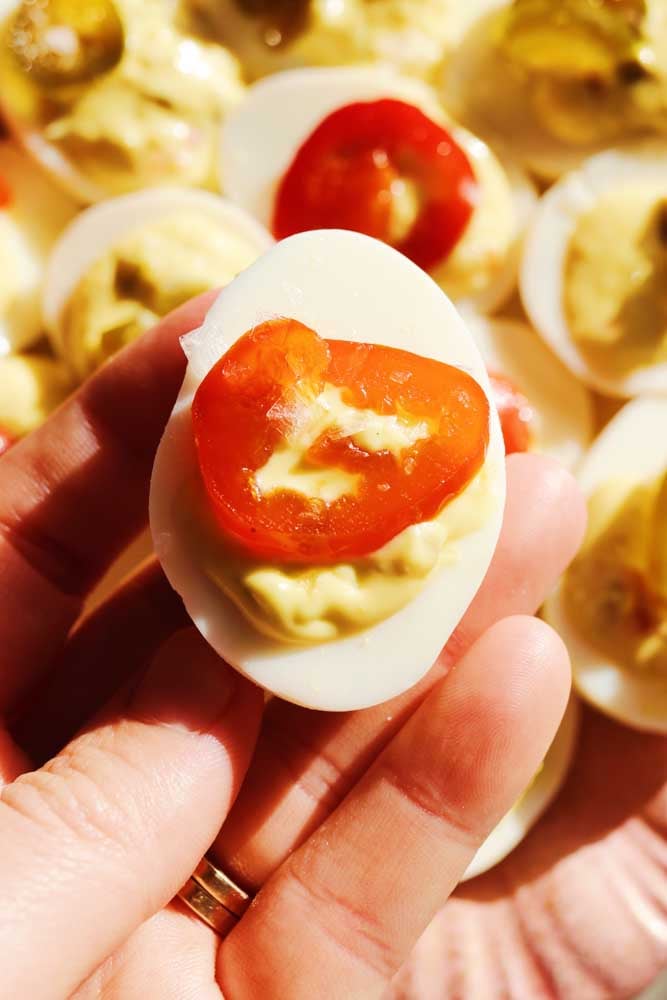 a hand holding a candied jalapeno deviled egg over a plate of them. 