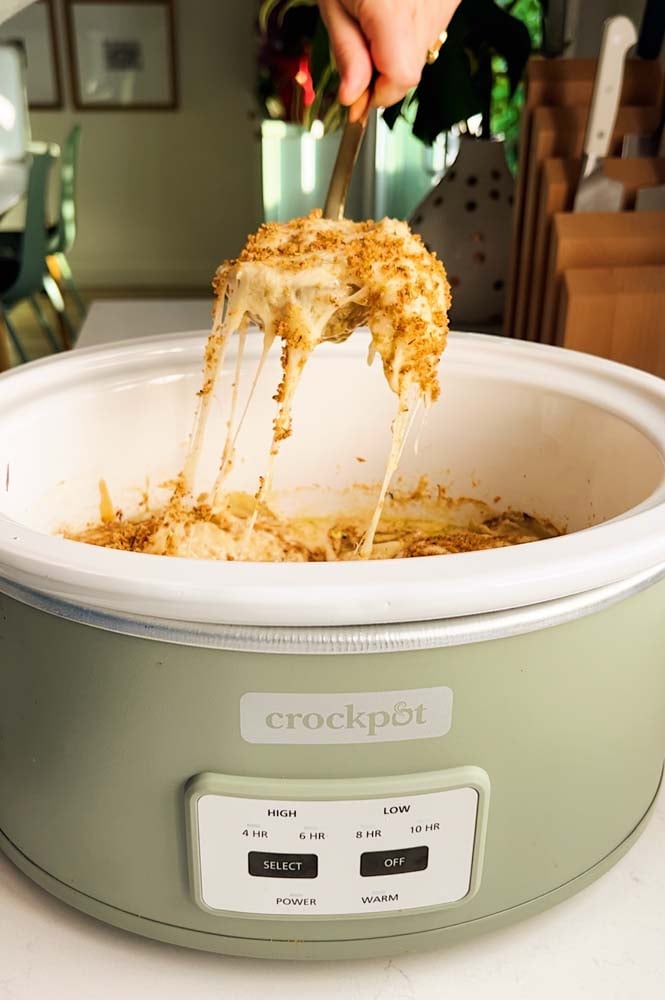 a green crockpot with a fork holding a scoop of cheesy crockpot potatoes au gratin with a glorious cheese pull!