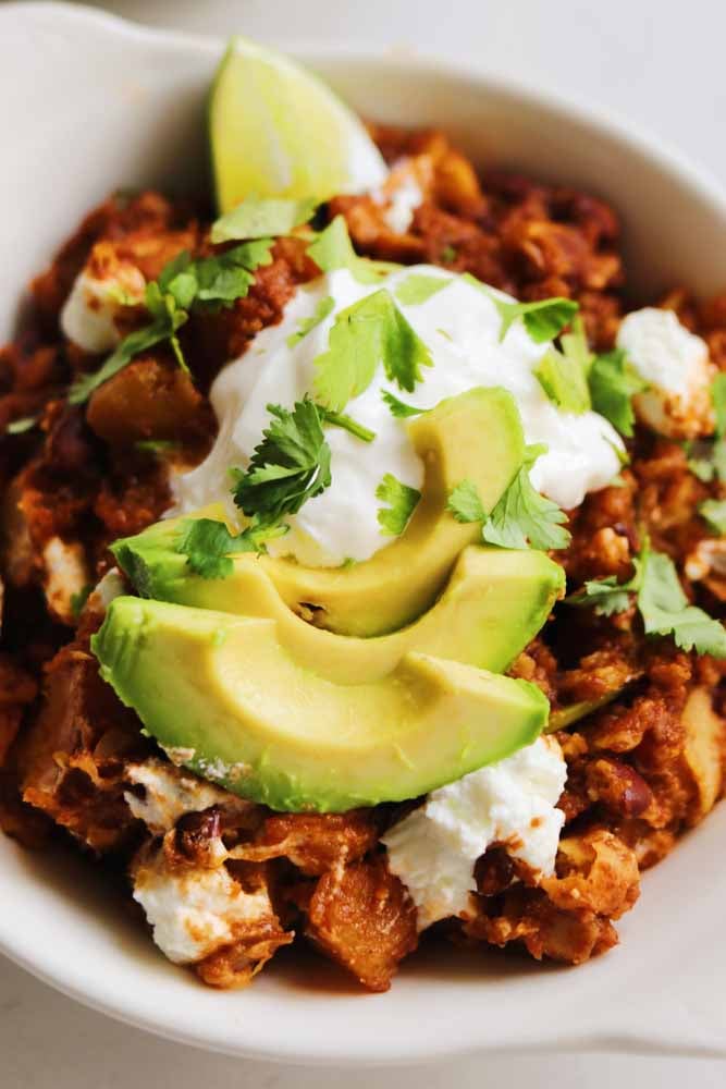 a bowl of crockpot enchilada casserole made with butternut squash and goat cheese with avocado and sour cream on top. 
