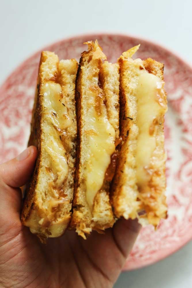 a hand holding 3 halves of a gruyere grilled cheese over a pink plate. 